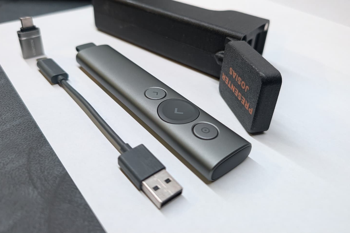 Logitech Spotlight Presenter mit USB C Adapter und kurzem Ladekabel vor dem schwarzen 3D gedruckten Case, dessen aufgeklappter Deckel orange mit PRESENTER JOSIAS beschriftet ist.