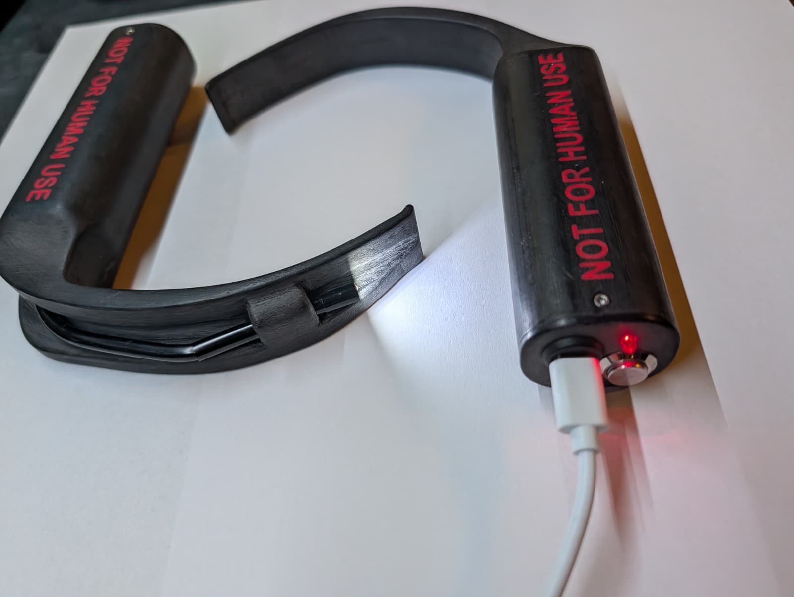 Zwei schwarze 3D gedruckte Videolaryngoskope mit roter Aufschrift NOT FOR HUMAN USE, links der hyperangulierte, rechts der Macintosh Spatel, der rechte hängt am USB C Ladekabel und leuchtet rot, die Spatelbeleuchtung ist eingeschaltet.