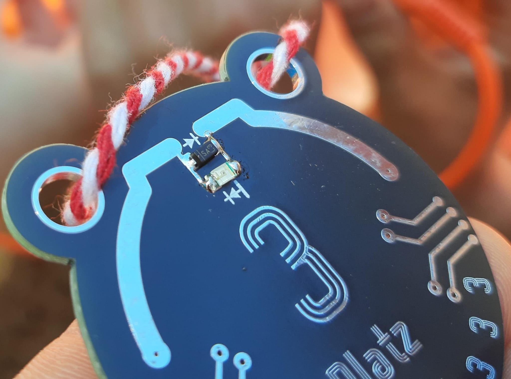 Nahaufnahme einer bärenförmigen Platinen-Medaille an roter Kordel, LED und Diode sind auf dem gefalteten Dipol in der Mitte verlötet, im Hintergrund ein unscharfes Lagerfeuer.